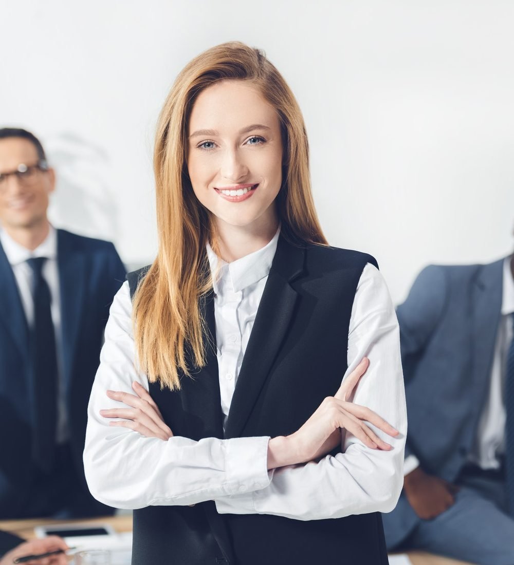 beautiful young businesswoman looking at camera with blurred colleagues on background e1647503302428
